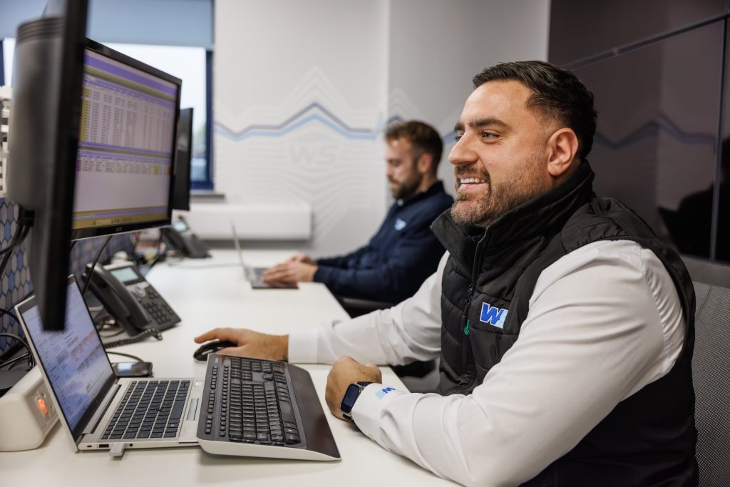 Warehouse worker at computer workstation reviewing logistics data on dual monitors, wearing high-visibility vest